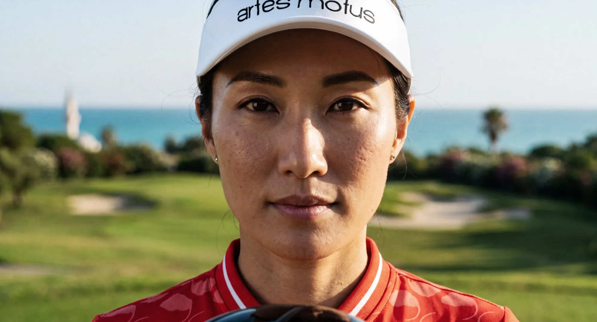 Portrait of the female golfer in a white Artes Motus visor and red top, fairway and sea behind her