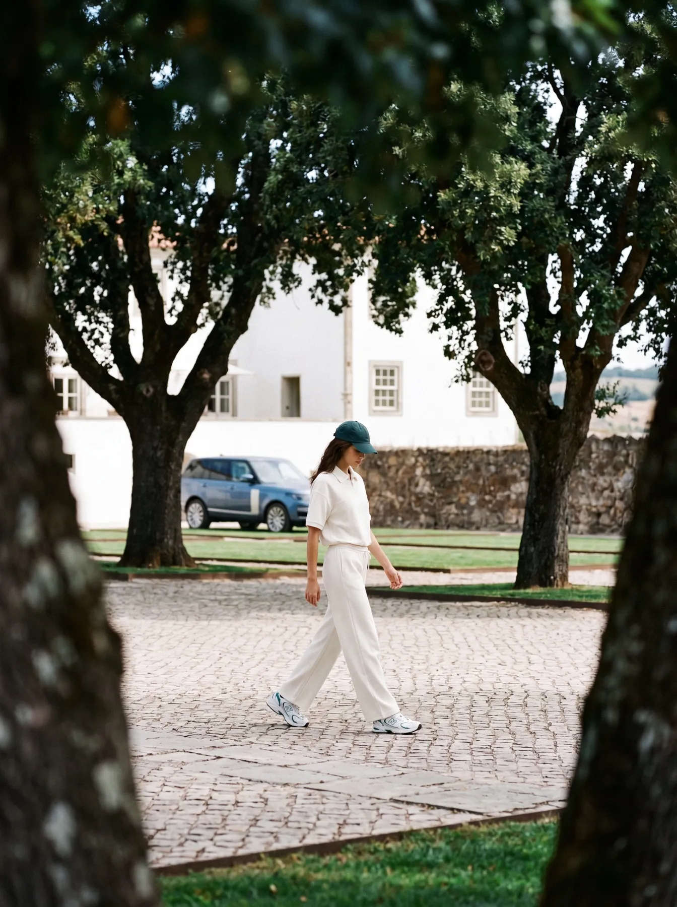 Model in a cream polo and trousers walking across the cobbled courtyard, a blue car parked behind the trees