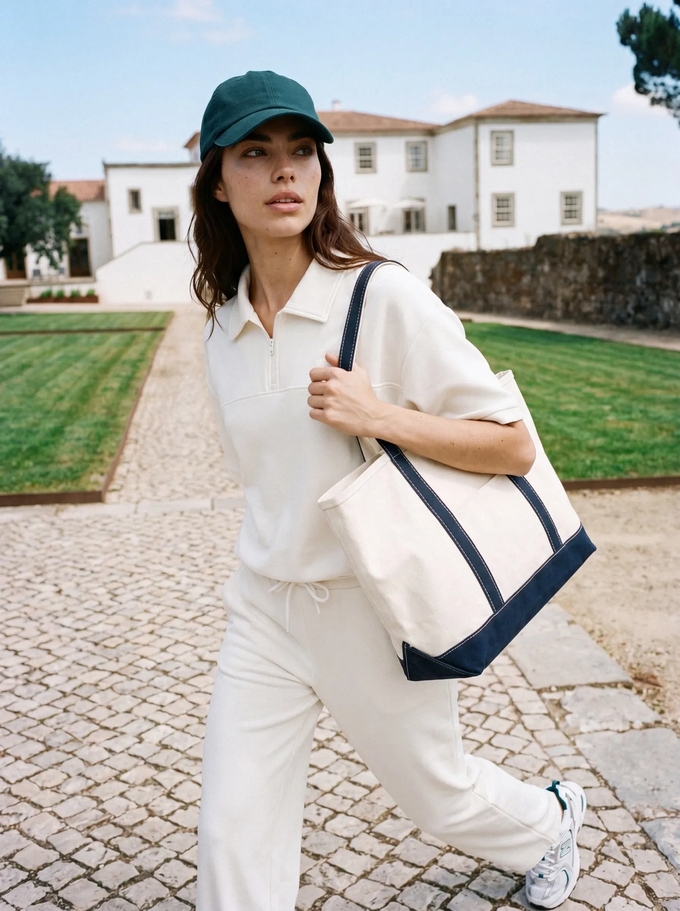 Model in a cream polo and a green cap carrying a canvas tote across a cobbled courtyard