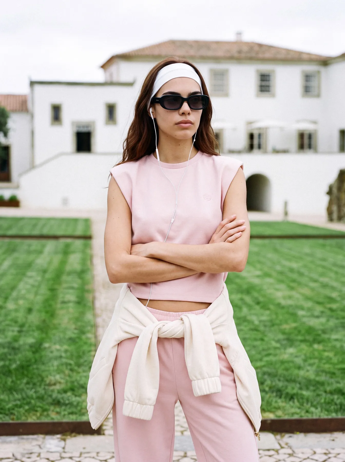 Model in a pink top and trousers with arms crossed on the courtyard lawn, the house behind her