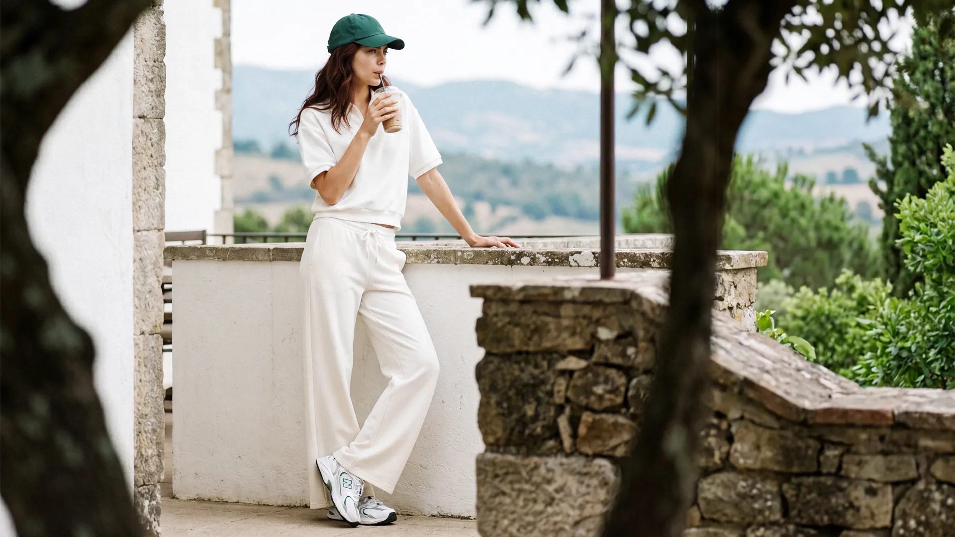 Model in a white top and trousers on the terrace, a drink in hand, the hills behind