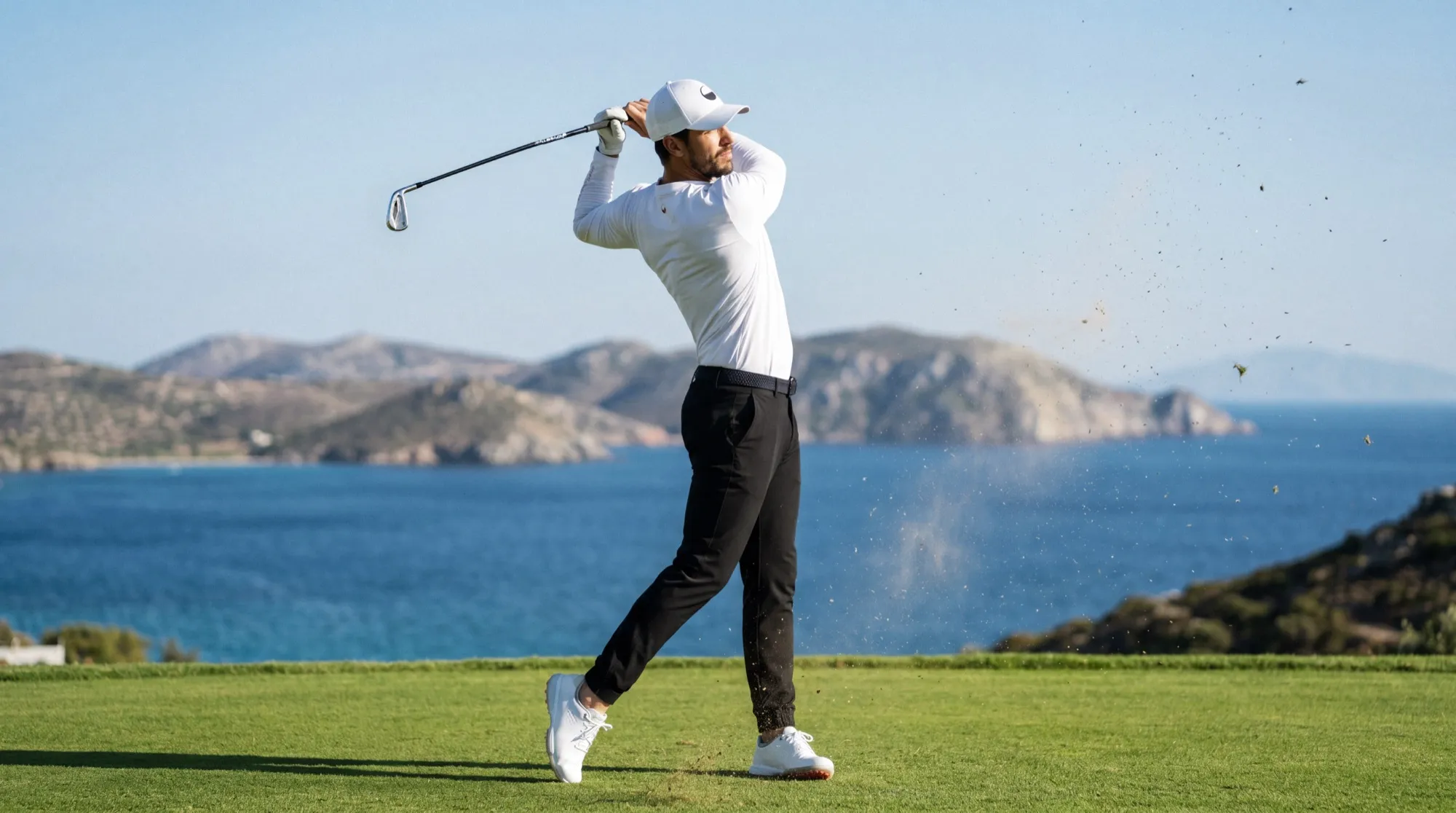 Male golfer in white and black holding his follow-through on a tee above a blue bay, turf flying
